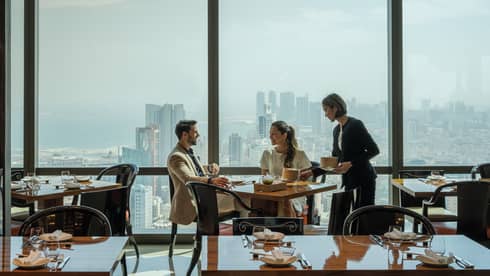 Several people meet in an Art Deco dining room overlooking the Bahrain cityscape. 