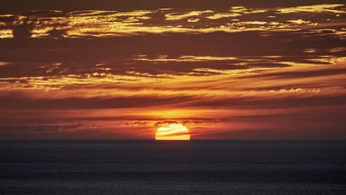 Dramatic yellow-and-orange streaked sky and dark, calm water below, top half of sun still visible in the distant horizon.