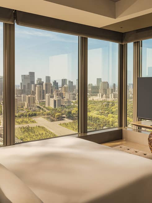 Luxury hotel room featuring king bed, TV and two walls of windows looking out to city and gardens