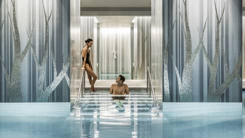 A couple enjoys a serene swimming pool, with one person seated on steps between elegant blue and silver columns.