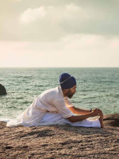 Man sits on sand beach near water, stretches his leg in a yoga pose 
