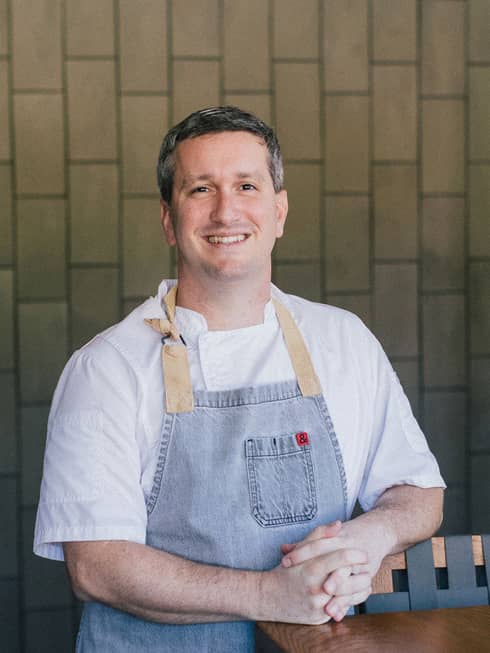 A man in a white shirt and blue apron leaning on a table.