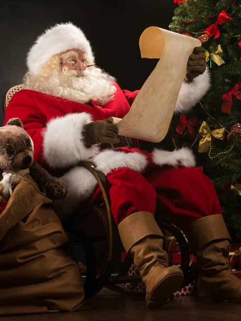 Santa Claus seated in large rocker holding a scroll, flanked by decorated Christmas tree with gifts and overflowing gift bag