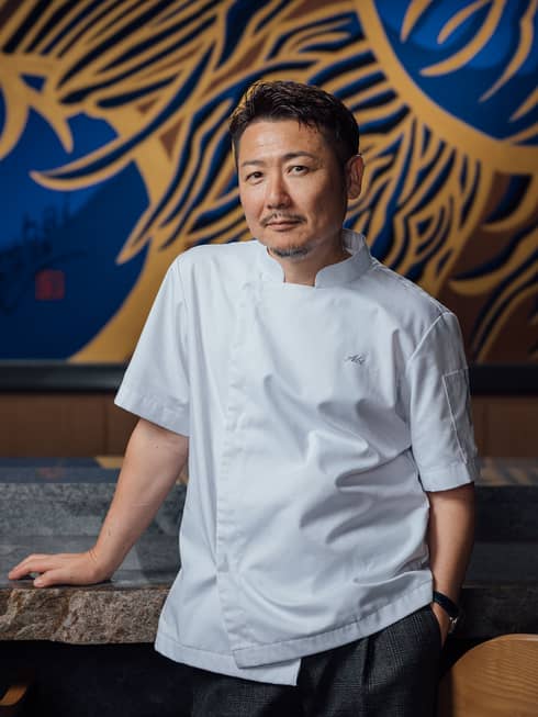 Chef with short dark hair and mustache wearing a white short-sleeve chef's jacket and black pants leans on a stone table with a gold-and-blue mural behind him