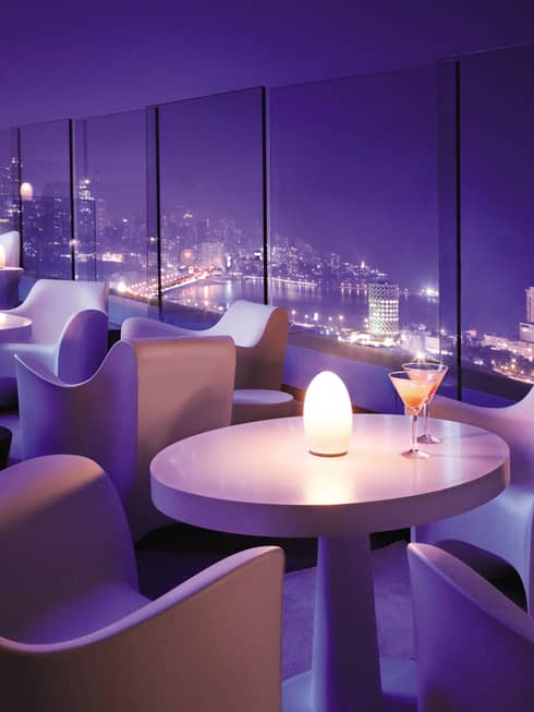 Modern white cocktail tables with candles in lounge with purple lighting, city night views