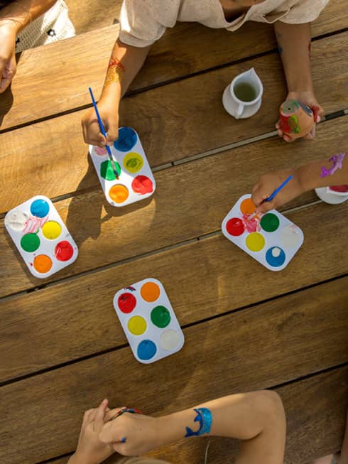 Overhead view of kids hands reaching into palettes of paint with their paint brushes on a wooden table