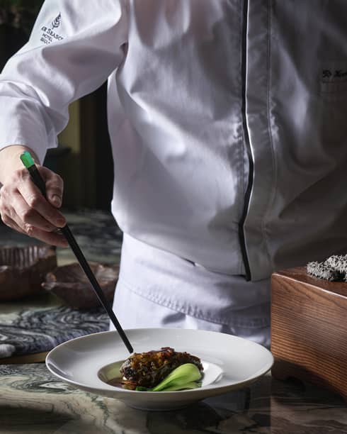 Chef using chopsticks to plate black pork belly in a white dish near a dozen black spiky delicacies on a small wood platform.