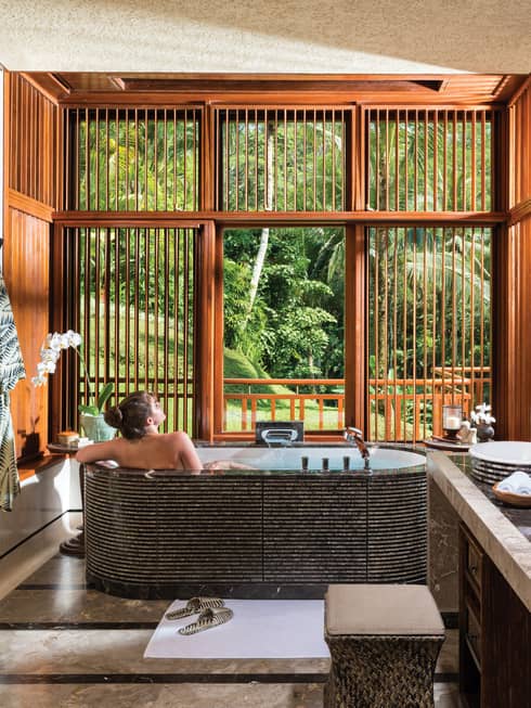 Woman with bare back relaxes in black freestanding spa tub near double sink, wood shutters open to lush garden view