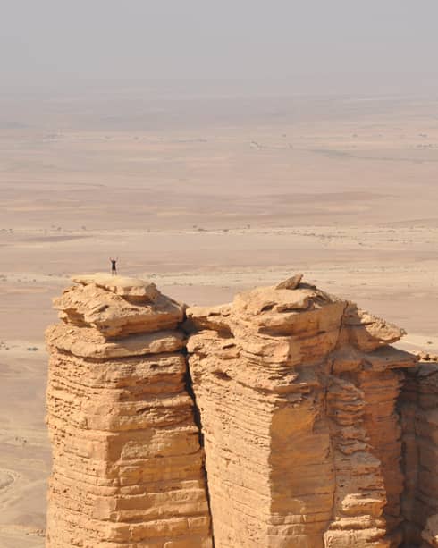 An aerial view of a giant golden cliff surrounded by a dry desert and a small silhouette of a person standing on top of it.
