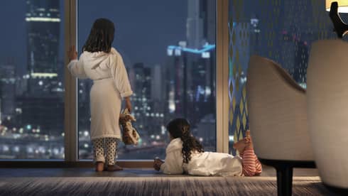 Two young children wearing bath robes look out a floor-to-ceiling window at a nighttime view of an urban cityscape.