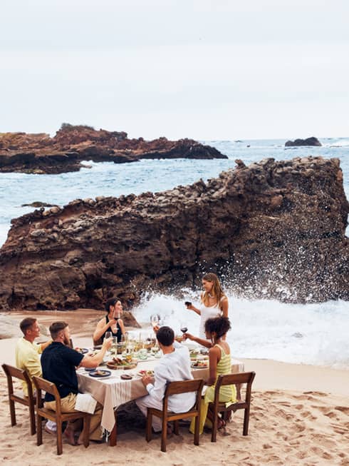 A group eating at a table on a beach with waves crashing nearby.