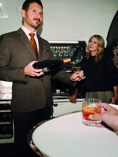 Person wearing brown tweed suit, white shirt and red tie pours a glass of wine for a private jet traveller standing nearby
