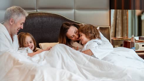 A family in bathrobes relaxing in a hotel bed under white blankets with a large upholstered headboard