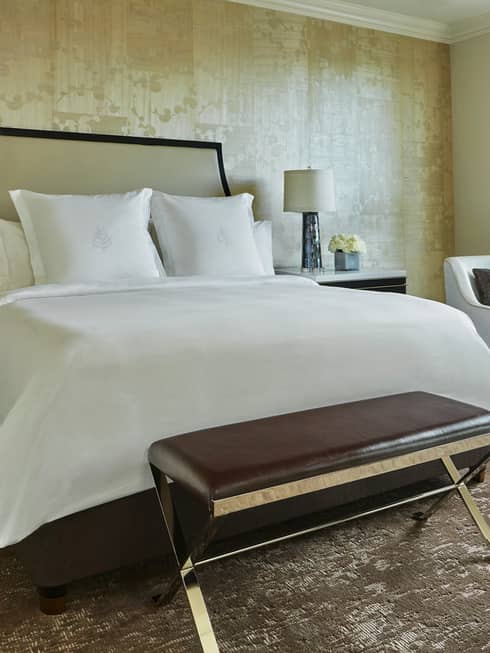 Georgetown suite with leather bench, bed with white linens, headboard against wallpaper, white armchair by sunny window