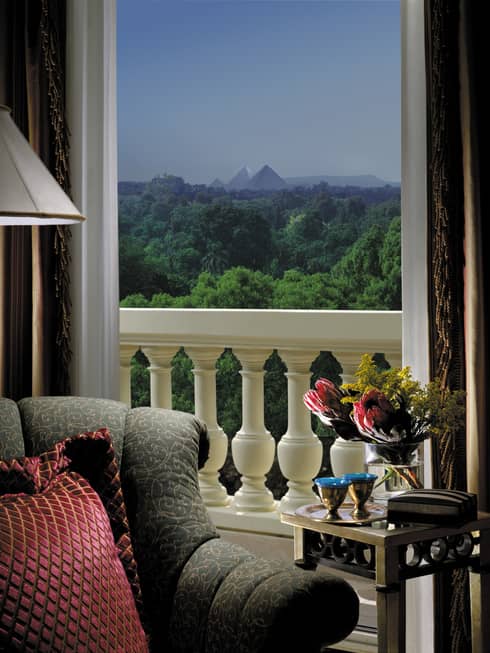 Close-up of plush armchair, red pillow, open patio door to balcony with white pillars with view of trees, pyramid