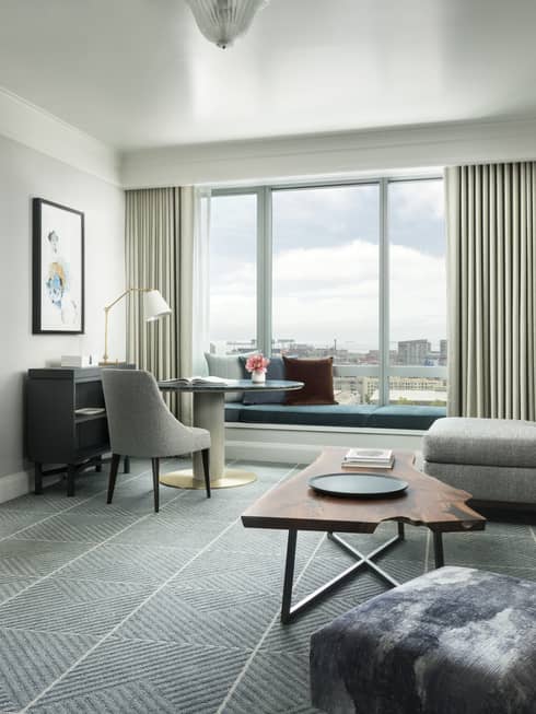 Hotel suite separate living room with retro-style L-shaped grey sofa and desk chair, live-edge wood coffee table