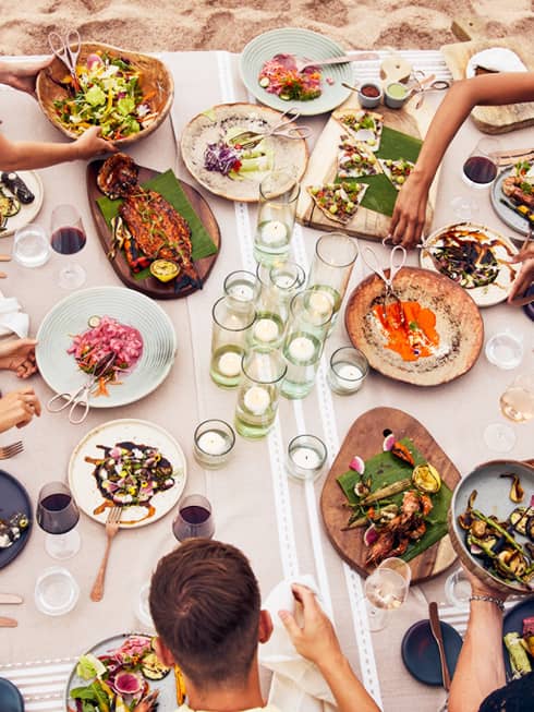 Beachside dining table with an assortment of vibrant dishes, candlelit glass holders and guests enjoying fresh seafood and grilled vegetables