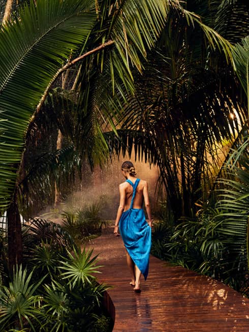 A woman in a blue dress walking along a wooden path covered in palm trees.