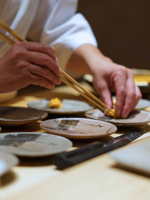 Chef carefully plating food with chopsticks on ceramic dishes in a Japanese restaurant setting.