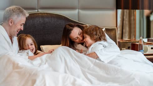 A family in bathrobes relaxing in a hotel bed under white blankets with a large upholstered headboard