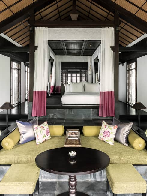 Elegant villa bedroom with a raised canopy bed surrounded by sheer curtains, overlooking a stylish seating area with a round table, cushioned sofas and large windows.