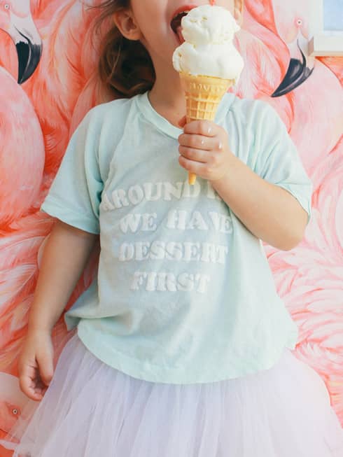 Child in front of a flamingo-motif wall with ice cream cone in one hand, wearing a tutu and t-shirt