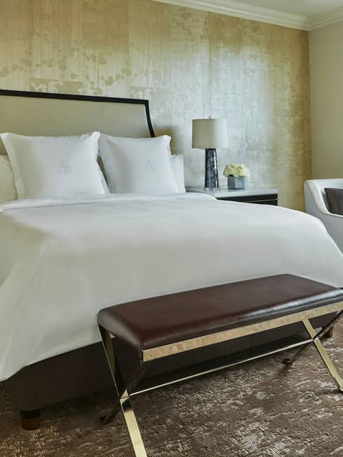 Georgetown suite with leather bench, bed with white linens, headboard against wallpaper, white armchair by sunny window