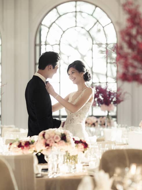 Woman in wedding dress adjusts groom's bow tie in light filled banquet room with pink flowers
