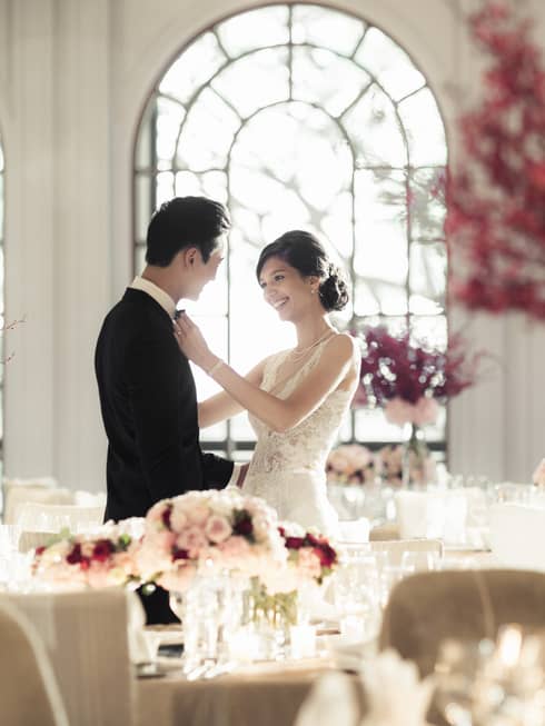 Woman in wedding dress adjusts groom's bow tie in light filled banquet room with pink flowers