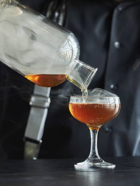 Smoke rolls off the decanter as a Four Seasons staff member pours bourbon into a coupe glass