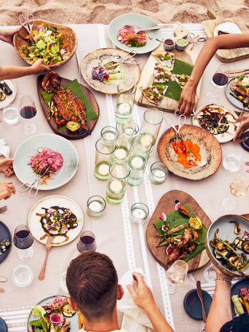 Beachside dining table with an assortment of vibrant dishes, candlelit glass holders and guests enjoying fresh seafood and grilled vegetables