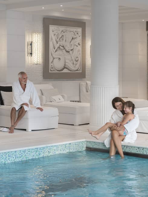 A child sits on her mother’s lap at the edge of an indoor pool as another guest smiles at them from a plush lounge chair.