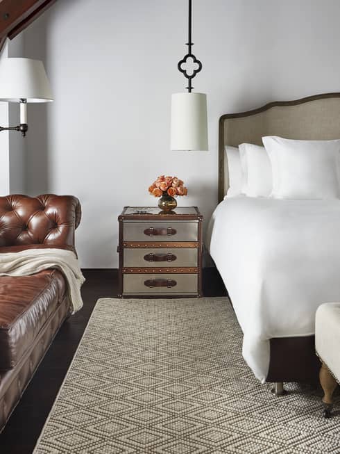 Brown period-style leather sofa under sloped beam roof next to nightstand, bed, cushioned bench