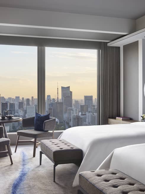 Guest room with two beds, tufted bench, seating area and floor-to-ceiling windows offering panoramic city views