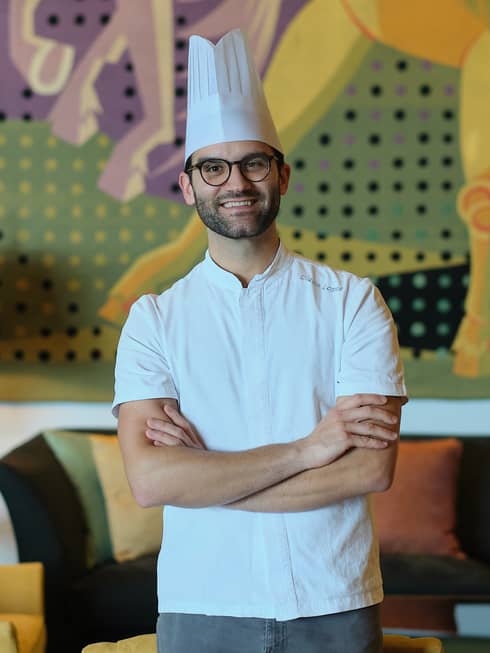 A chef in a white shirt and toque stands with arms crossed in a cozy interior space with a table lamp, upholstered chair and couch, and a mural on the back wall