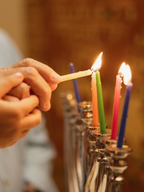 Two hands light multi-coloured candles on a menorah