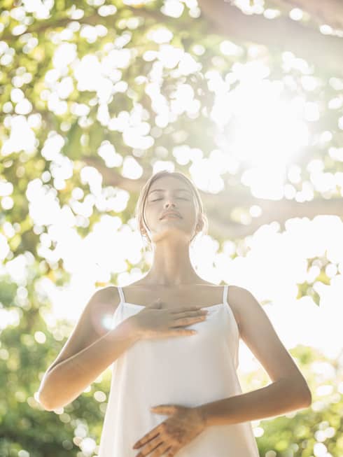 Person practicing mindful breathing in a wellness haven, standing under sunlit trees with hands on chest and abdomen