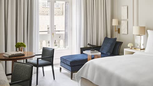Luxury hotel bedroom with a king bed, tufted headboard, blue chaise lounge, round table with books and French doors with balcony view