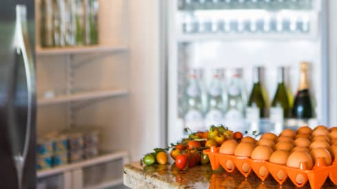 Smiling man holds green bottle in front of open fridge, fresh eggs on counter