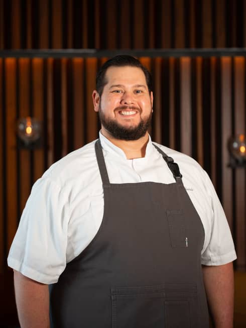 A male chef in a dark grey apron and white chef clothes with candles behind him.