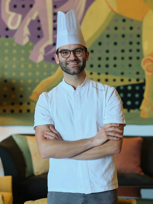 A chef in a white shirt and toque stands with arms crossed in a cozy interior space with a table lamp, upholstered chair and couch, and a mural on the back wall