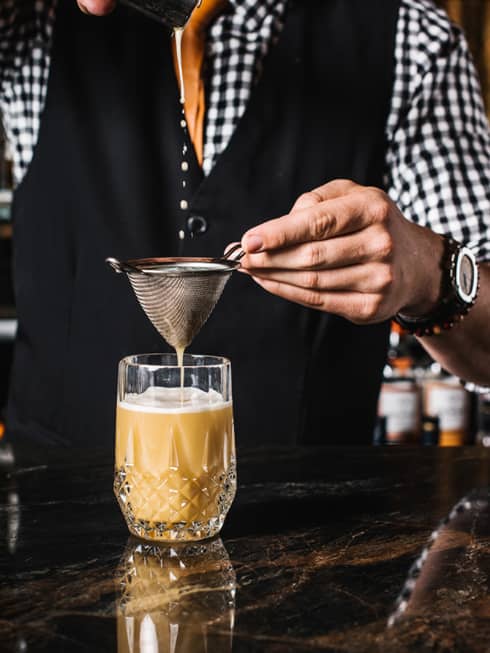 Grain bartender in vest, checkered shirt strains cocktail into crystal glass