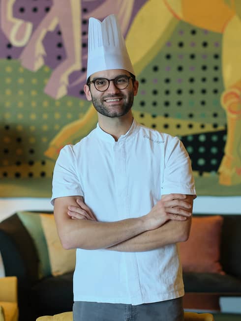 A chef in a white shirt and toque stands with arms crossed in a cozy interior space with a table lamp, upholstered chair and couch, and a mural on the back wall