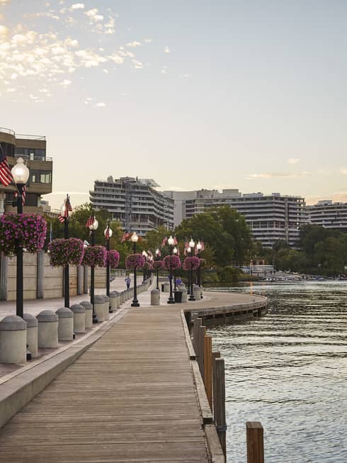 A waterfront with a wooden walkway next to light posts and flowers with building on the other side of a small road.