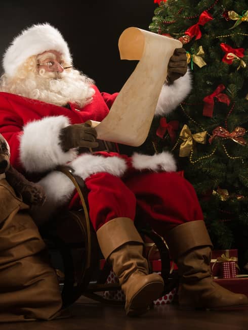 Santa Claus seated in large rocker holding a scroll, flanked by decorated Christmas tree with gifts and overflowing gift bag