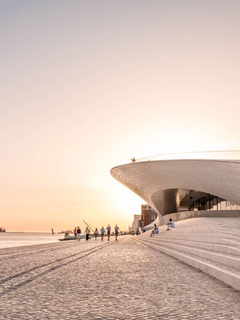 Modern waterfront building with sweeping architectural curves, people walking along a cobblestone promenade at sunset.