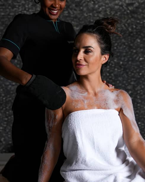 Woman with dark hair tied in a bun at the top of her head and wearing a white towel wrapped around her body is having her shoulders scrubbed by a spa attendant using a exfoliation glove