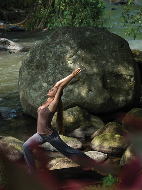 A guest practices yoga on the banks of a forest river; sunlight dapples the flowing water and the large, mossy boulders.