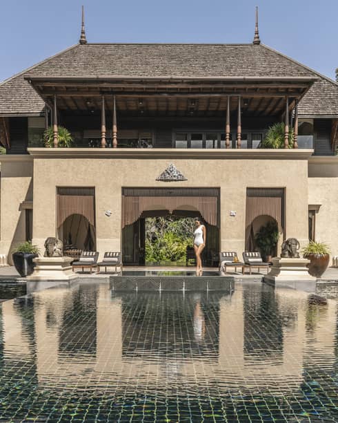 Person walking along the edge of a large reflective pool in front of a grand villa with traditional architecture surrounded by trees and outdoor seating