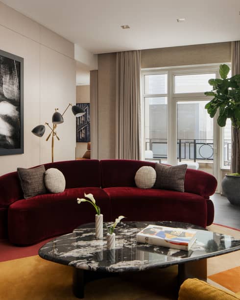 Luxury suite living room with burgundy sofa, coffee table, two yellow armchairs, large black-and-white wall art, potted plant and city-view balcony
